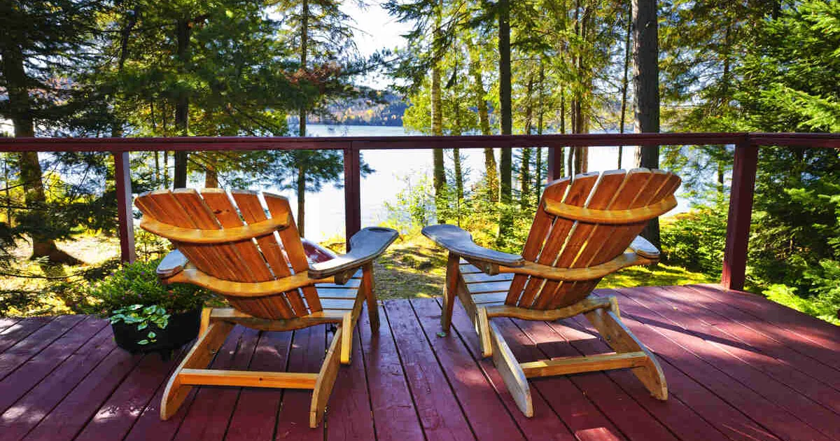 Two empty Muskoka chairs sit on a cottage deck overlooking a serene lake through a cluster of tall pine trees.