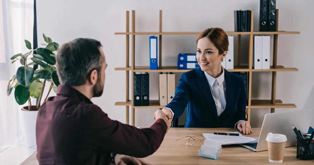 A female lawyer in a blue suit engaging in a handshake with a man in a burgundy shirt. Set in a bright office with a wooden table, potted plant, and organized shelves, it conveys a positive, collaborative mood ideal for business, meetings, or successful negotiations.