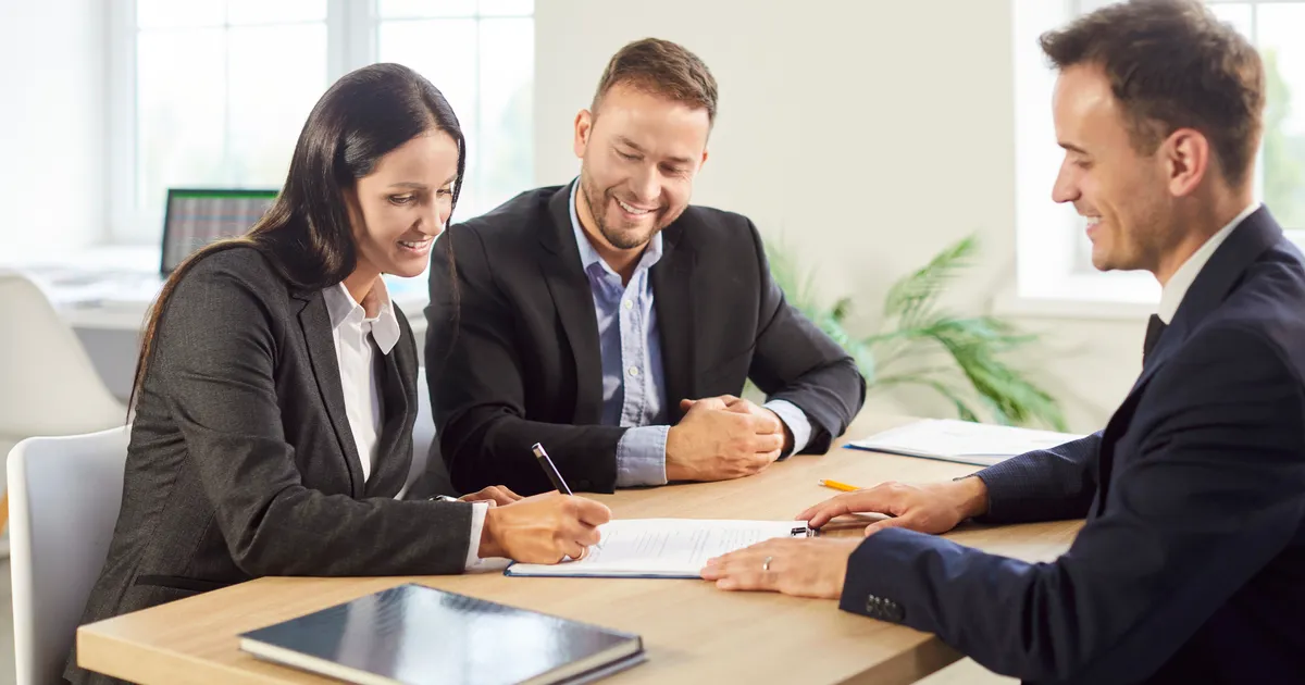 Three people in business attire smile around a wooden table in a bright office while one of them signs a document.