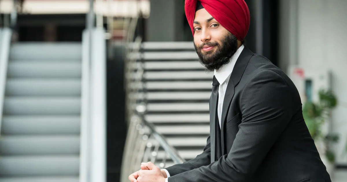 A well-dressed man in a bright red turban leans against a railing inside an office building.