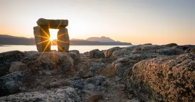 The setting sun shining through an inuksuk on a rocky shore above the fjord at Qikiqtarjuaq, Nunavut